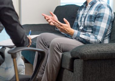 Man talking and waving hands in therapy session with psychiatrist, psychologist, counselor, therapist or life coach. Meeting with professional specialist with a notepad. Male patient sitting on couch.