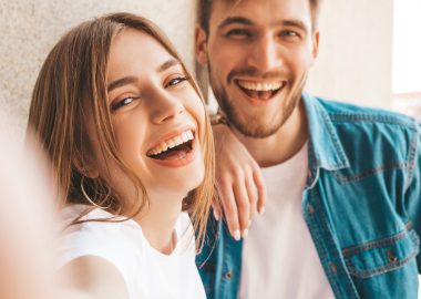 Smiling beautiful girl and her handsome boyfriend in casual summer clothes. Happy family taking selfie self portrait of themselves on smartphone camera. Having fun on the street background