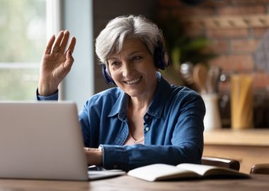 Close up smiling mature grey haired woman wearing headphones making video call, waving hand at webcam, greeting, chatting with relatives or friends, sitting at desk, looking at laptop screen